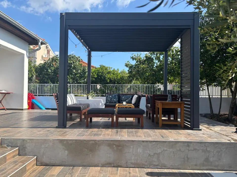 Covered lounge terrace at Villa Marco Polo with sofas under a slatted pergola, olive trees behind